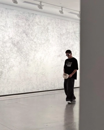 Man in casual clothing walking in a minimalist art gallery, gazing down. Neutral colors, with a textured wall and modern lighting.
