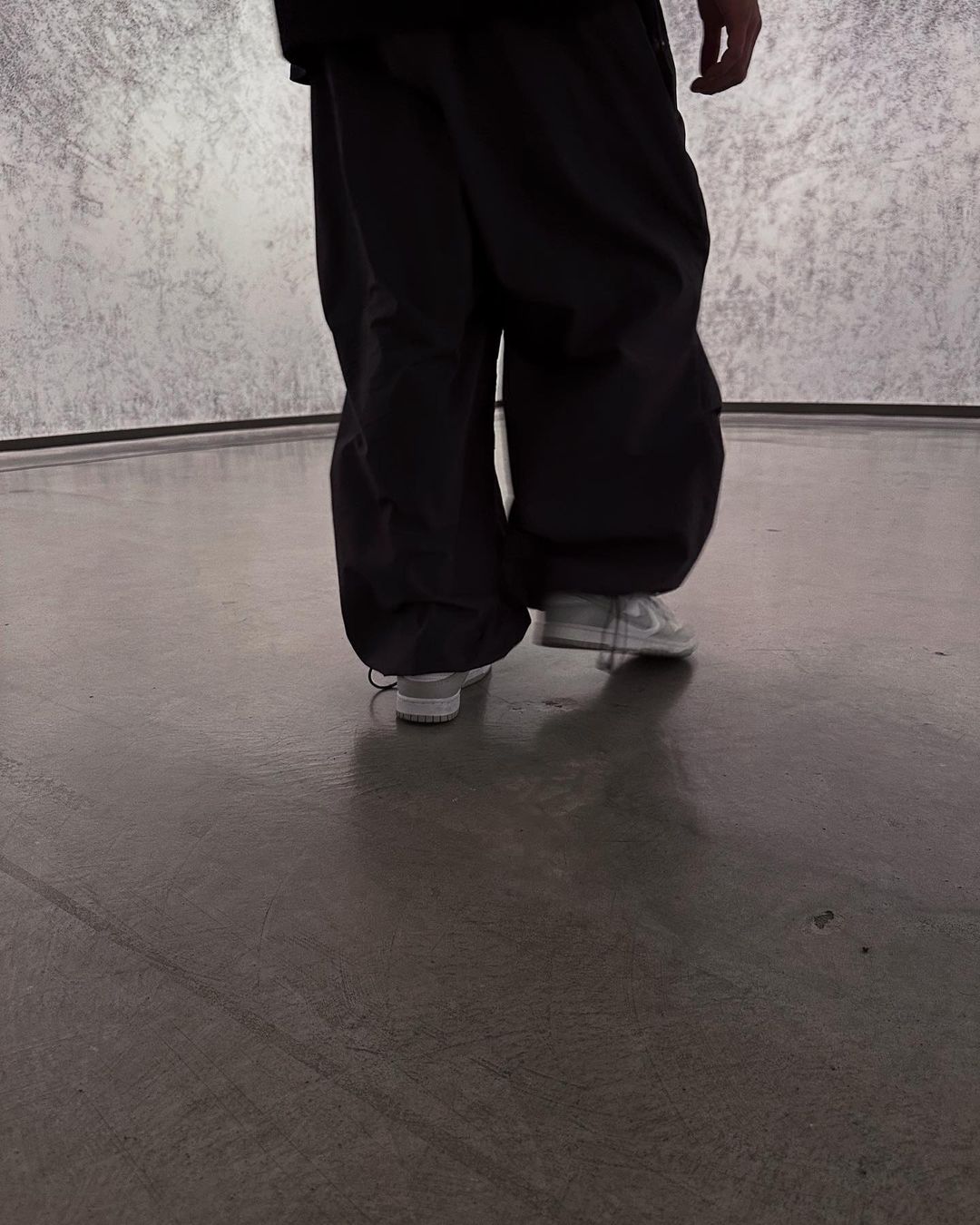 Person in baggy black pants and white sneakers walking on a smooth grey surface with a mottled grey wall in the background.