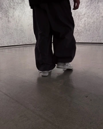 Person in baggy black pants and white sneakers walking on a smooth grey surface with a mottled grey wall in the background.