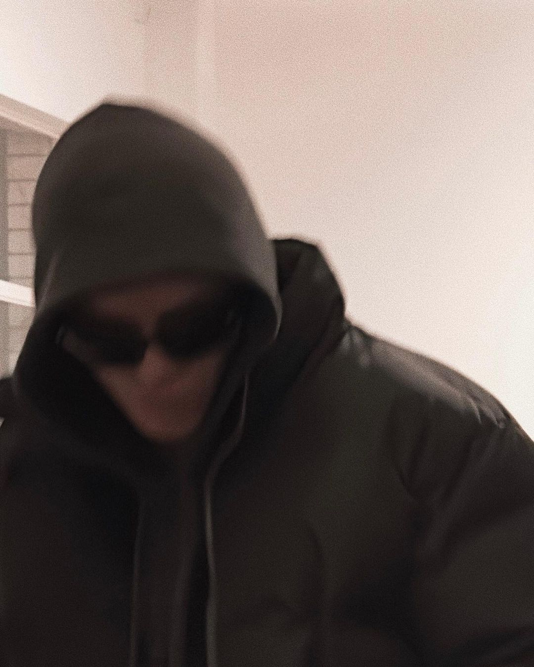 An adult wearing a dark puffer jacket with a hood and sunglasses, photographed indoors with neutral lighting and a slightly blurred effect.