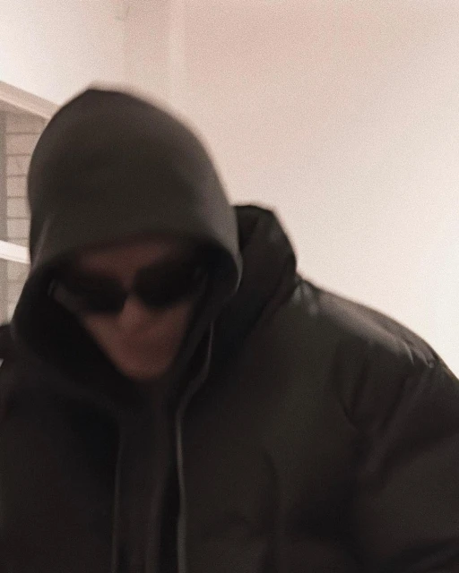 An adult wearing a dark puffer jacket with a hood and sunglasses, photographed indoors with neutral lighting and a slightly blurred effect.