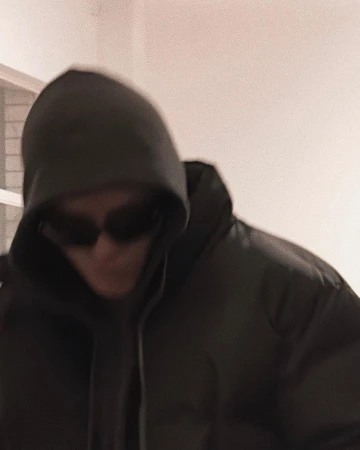 An adult wearing a dark puffer jacket with a hood and sunglasses, photographed indoors with neutral lighting and a slightly blurred effect.