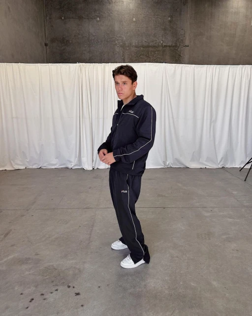 Young man in a dark blue Fila tracksuit standing indoors in front of a white curtain and concrete wall. Fashion shot in studio.