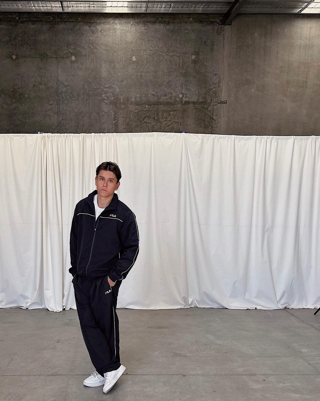 Adult male in a dark blue FILA tracksuit and white sneakers, standing against a white curtain backdrop in an indoor industrial setting.