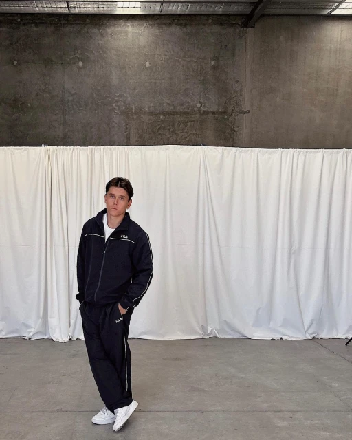 Adult male in a dark blue FILA tracksuit and white sneakers, standing against a white curtain backdrop in an indoor industrial setting.