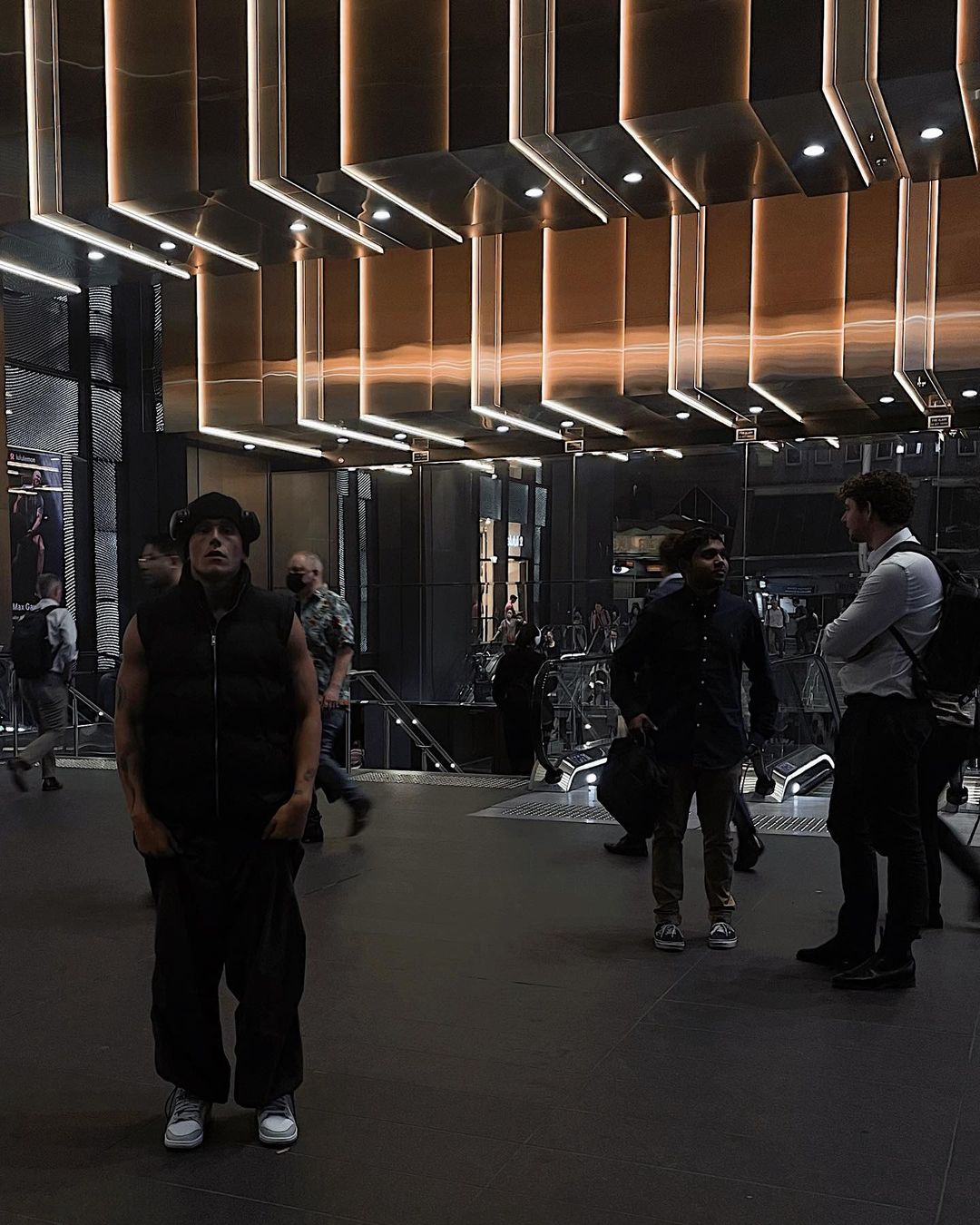Man stands in a modern train station, wearing a black vest, with other people walking in the background and illuminated ceiling.