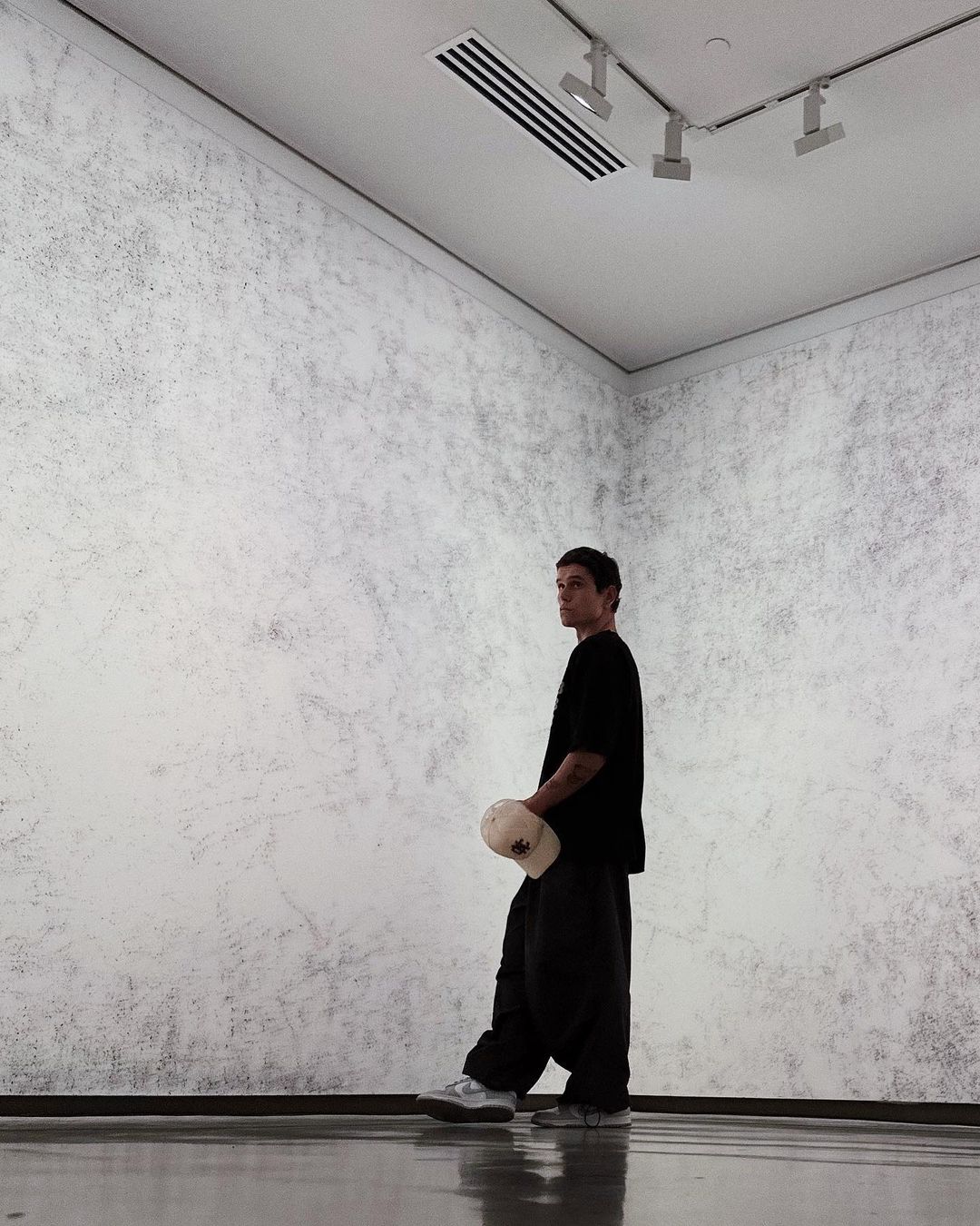 A young man in a black t-shirt and baggy pants standing in a minimalist art gallery with gray walls holding a cap.