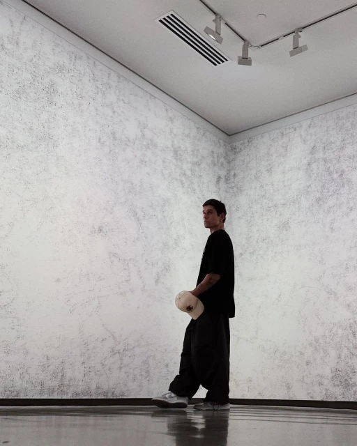 A young man in a black t-shirt and baggy pants standing in a minimalist art gallery with gray walls holding a cap.