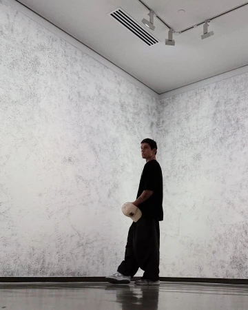 A young man in a black t-shirt and baggy pants standing in a minimalist art gallery with gray walls holding a cap.