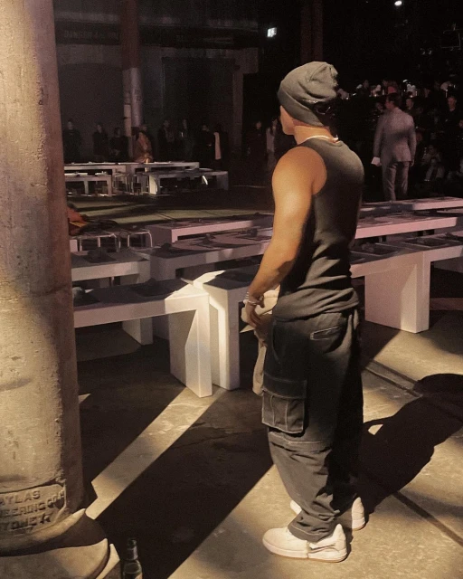 Man stands in fashion show venue wearing a tank top, cargo pants, beanie. A blurred crowd fills the background; rows of tables in foreground.