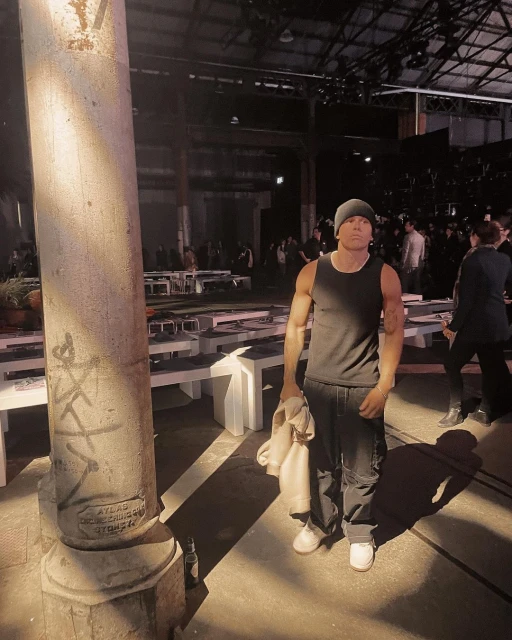 Man stands in a tank top and beanie at fashion show. Empty white benches and a crowd are visible in background. 