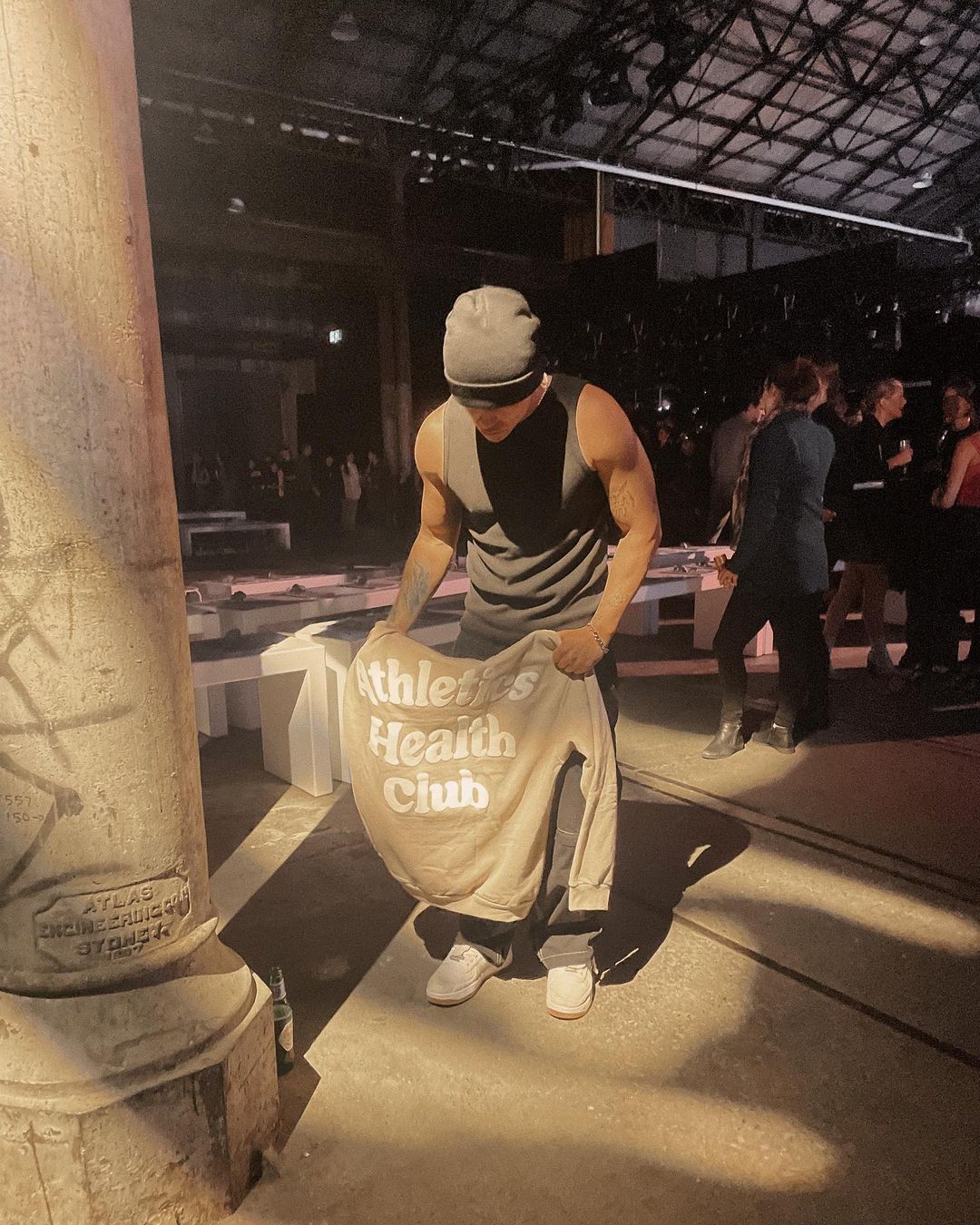 Adult male holds a beige sweatshirt with ‘Athletics Health Club’ text in an indoor space with concrete columns and a crowd of people.