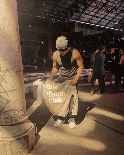 Adult male holds a beige sweatshirt with ‘Athletics Health Club’ text in an indoor space with concrete columns and a crowd of people.