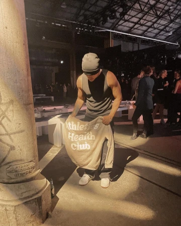 Adult male holds a beige sweatshirt with ‘Athletics Health Club’ text in an indoor space with concrete columns and a crowd of people.