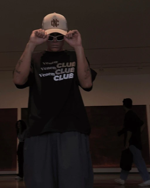 A young adult wears a baseball cap and sunglasses indoors, showcasing a graphic T-shirt, with people and artworks in the background.