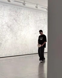 Man in black t-shirt and dark baggy trousers wearing white sunglasses standing in a gallery interior.