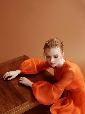 A woman in an orange dress leans on a wooden table, set against a neutral backdrop in a fashion editorial style photograph.