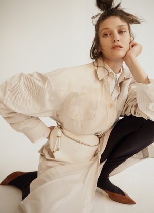 Woman in beige jacket poses in a photo studio, wearing black leggings and brown shoes, with neutral background and dramatic fashion style.