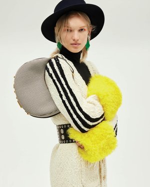 Fashionable woman in studio wearing hat, oversized sweater with stripes, green earrings, and studded belt against a white background.