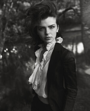 Black and white portrait of a young woman in a blazer and ruffled top, standing against a blurred background of trees in an outdoor setting.