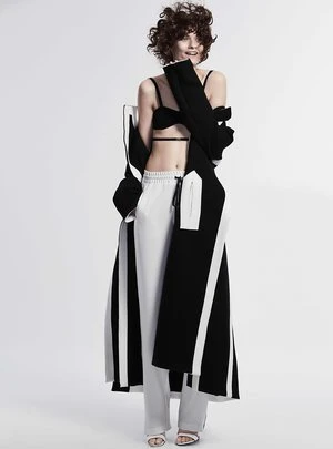 Model with curly dark hair wearing a black bralette, white trousers, and an open black and white coat against a plain background