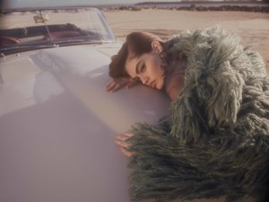 Female model in a fuzzy coat poses with her head on the hood of a vintage white convertible in a desert environment on a sunny day.