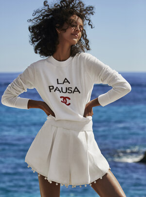 A woman in a white long-sleeved shirt with the words 'La Pausa' and a red logo paired with a white pleated skirt smiles on a sunny day.