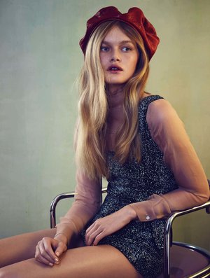 A fair-skinned adult female in a red leather beret poses in a sparkly dress with sheer sleeves while seated in a metal chair.