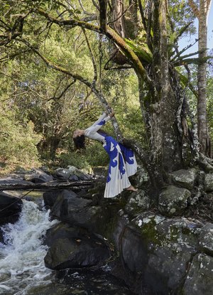 Woman in white and blue dress bending backward under a tree branch near a rocky river in an outdoor forest environment.