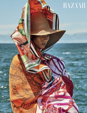 Woman wearing a large hat and patterned scarf in an outdoor fashion shot, sea in the background, 'BAZAAR' logo visible.