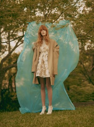 A woman poses outdoors in a floral dress and trench coat against a blue plastic backdrop with green grass and trees surrounding.