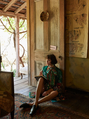 Woman in a floral dress sits on a rug, writing in a notebook, posed indoors near an open door with natural light and vintage aesthetics.