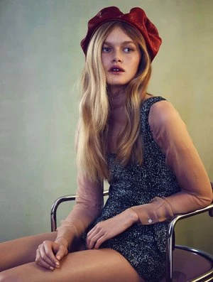 Model wearing a red beret and textured dark romper seated on a metallic chair against a muted green wall.