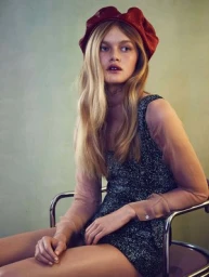 Model wearing a red beret and textured dark romper seated on a metallic chair against a muted green wall.