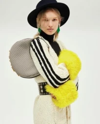 Model wearing a black hat, cream textured cardigan, and holding a structured handbag against a plain background