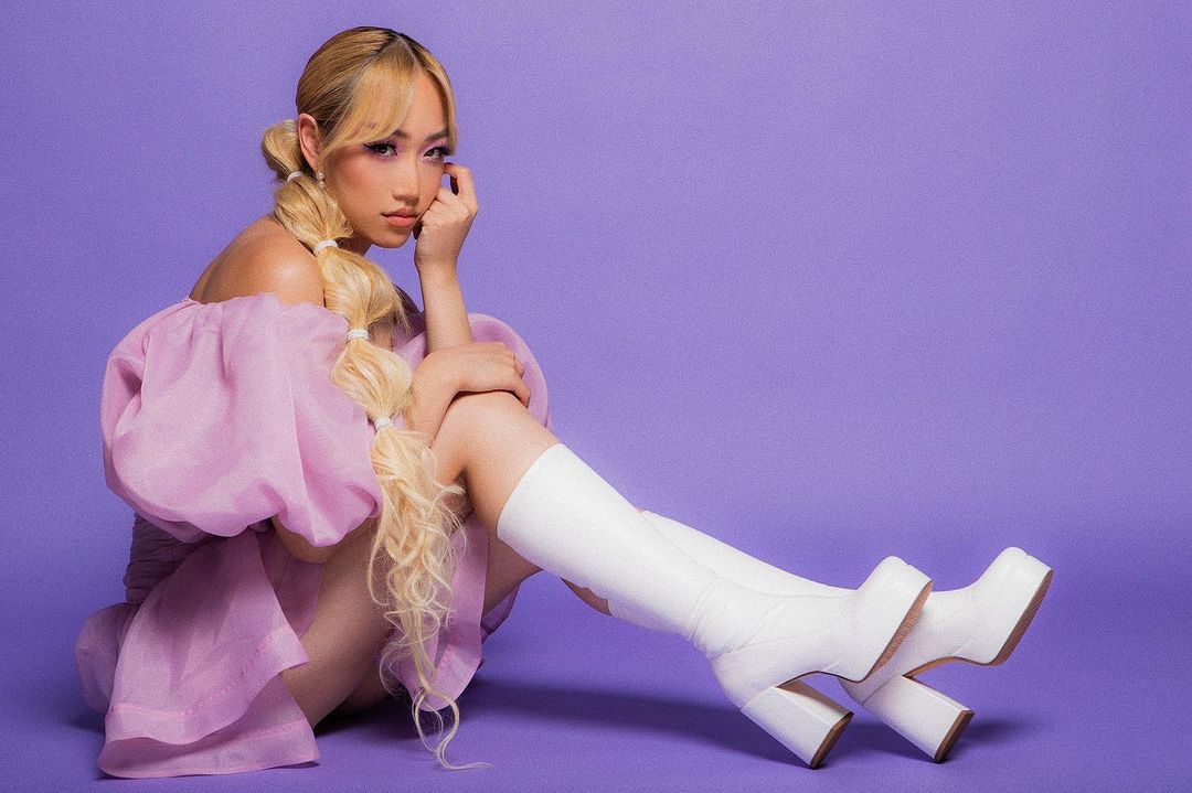 Portrait of a blonde woman in a lilac dress and white boots sitting against a purple background in a studio with soft, diffused lighting.