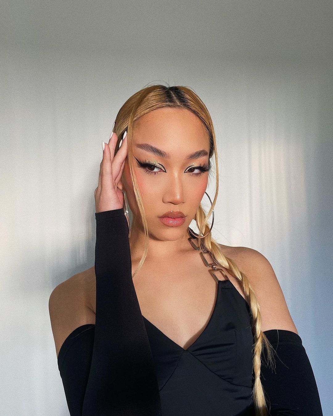 Woman with blonde hair and dramatic eye makeup posing in front of a white wall, wearing a black top and hoop earrings.