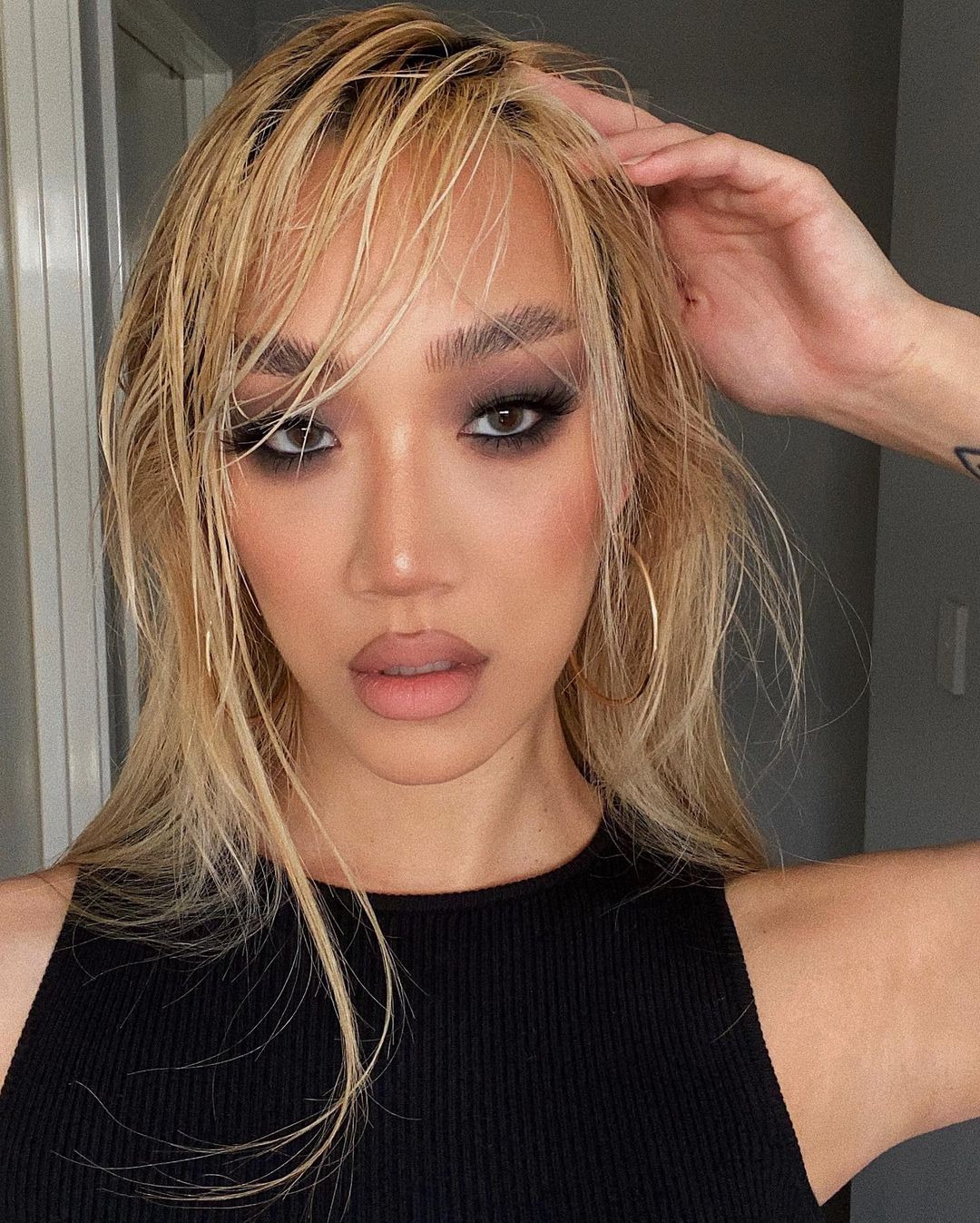 Close-up of a young adult woman with blonde hair, dark eye makeup, and gold hoop earrings wearing a black tank top indoors.