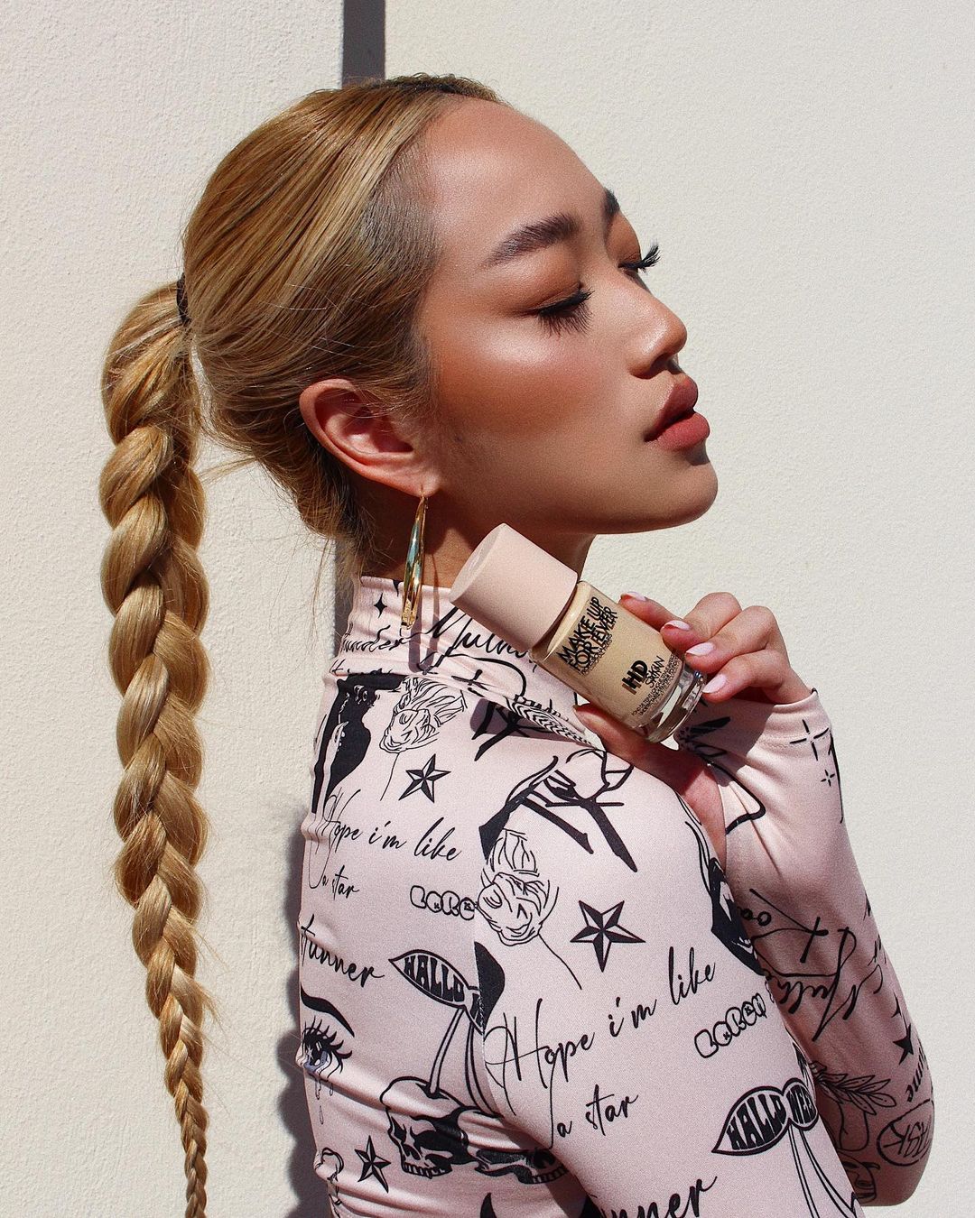 Profile of an adult woman with blonde hair in a ponytail holding a makeup foundation bottle while wearing patterned clothing.