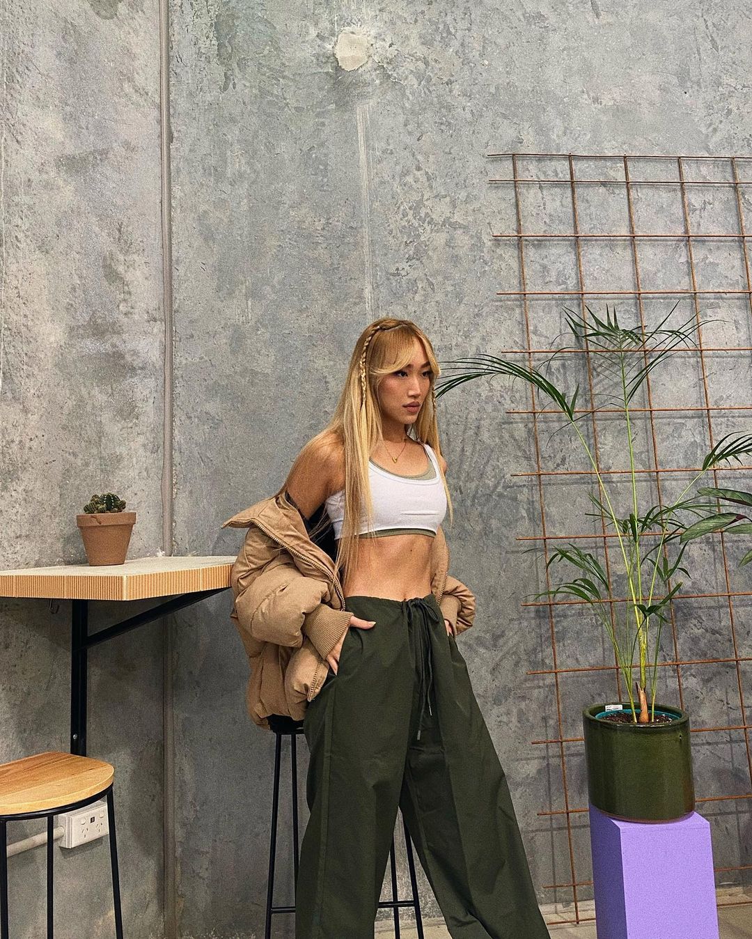 A young woman in athletic wear and puffy jacket poses in an indoor setting with a concrete wall and plant behind her, styled casually.