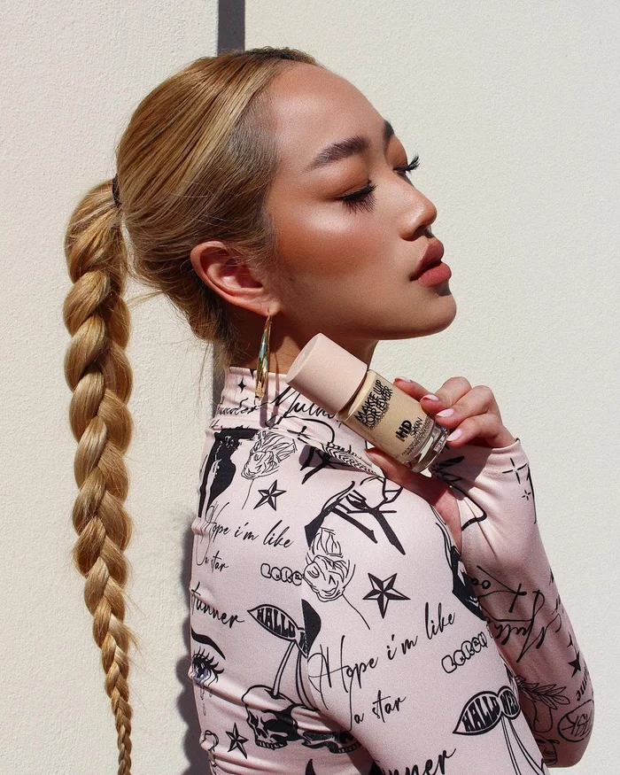 Close-up profile of a woman with long blonde braided hair holding a small cosmetic bottle against a textured wall.