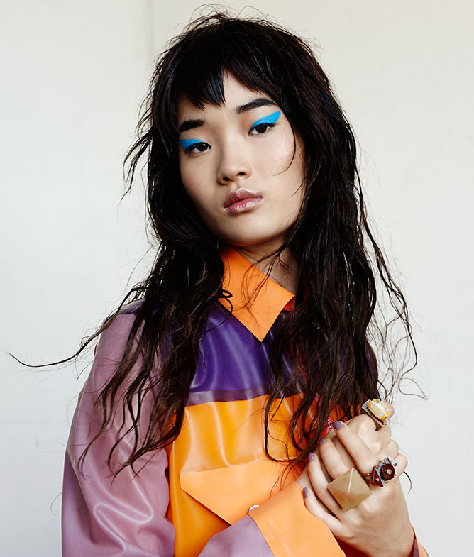 Portrait of an Asian woman with turquoise eyeshadow wearing a modern color-blocked shirt and sculptural rings against a white background.