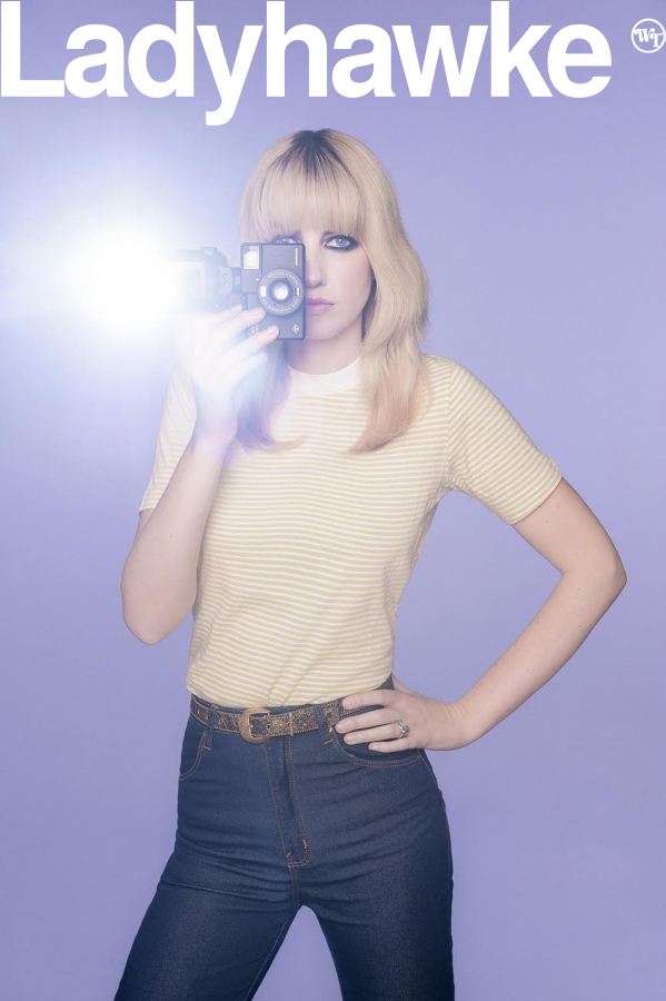 Woman holding vintage camera with flash, wearing striped shirt and blue jeans against lilac background, album cover art.