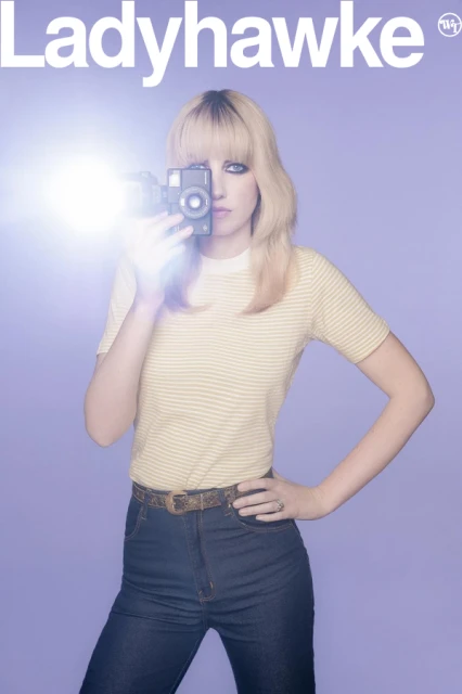 Woman holding vintage camera with flash, wearing striped shirt and blue jeans against lilac background, album cover art.