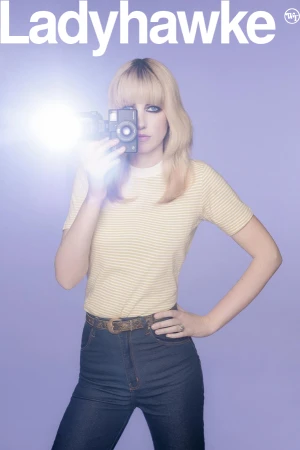 Woman holding vintage camera with flash, wearing striped shirt and blue jeans against lilac background, album cover art.