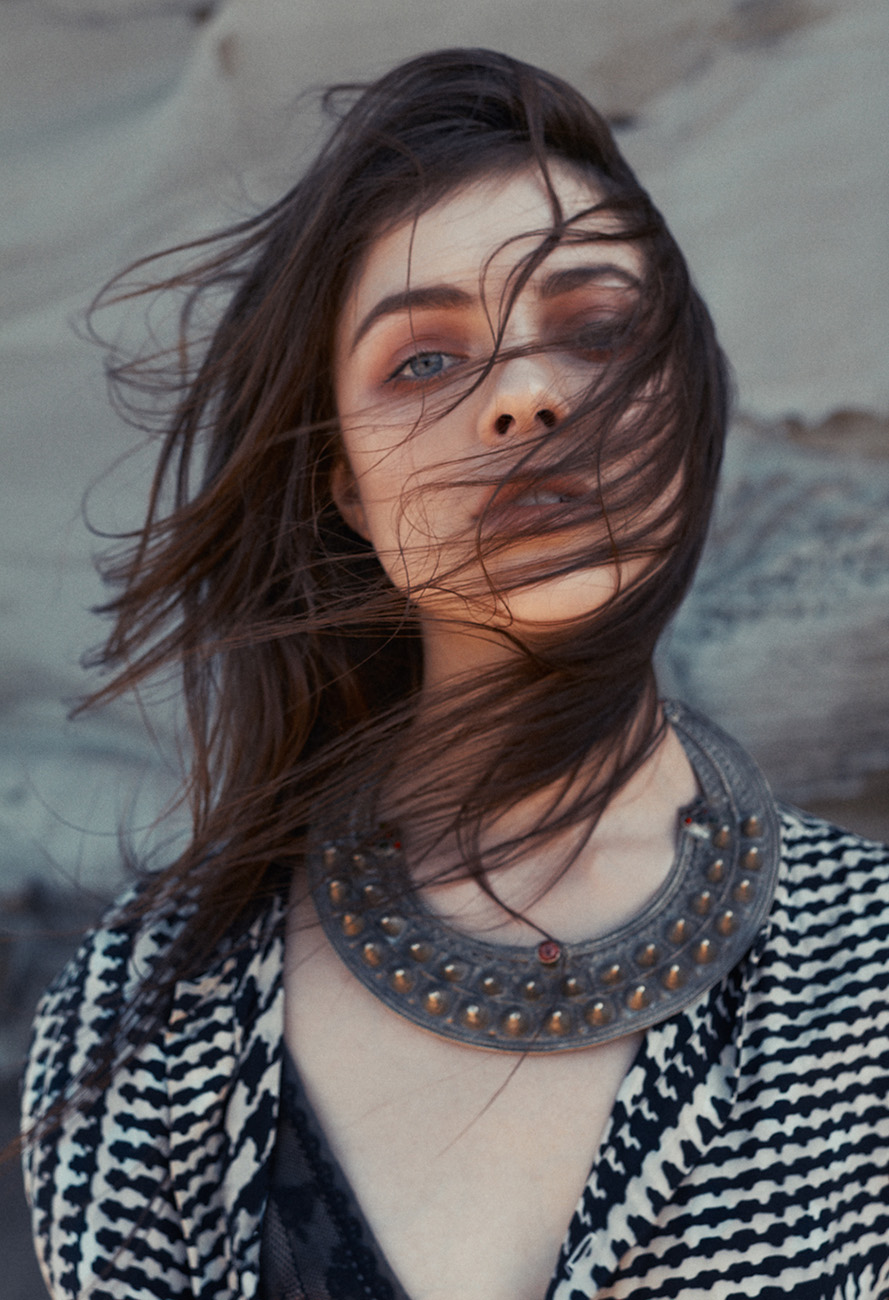 Eye-level portrait of a female adult with blue eyes and wind-blown dark hair, wearing a patterned jacket and a large ornamental necklace.