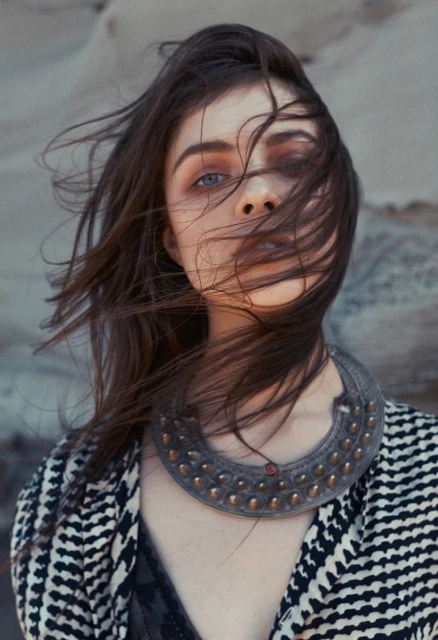 Eye-level portrait of a female adult with blue eyes and wind-blown dark hair, wearing a patterned jacket and a large ornamental necklace.