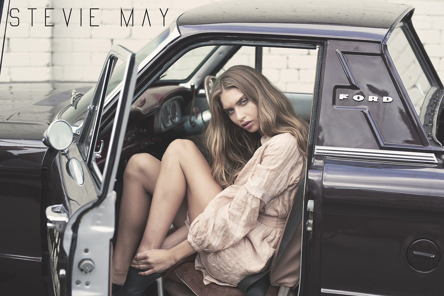 Young woman with long brown hair posing in a vintage black Ford car wearing a pale pink dress with an open door and white brick background.