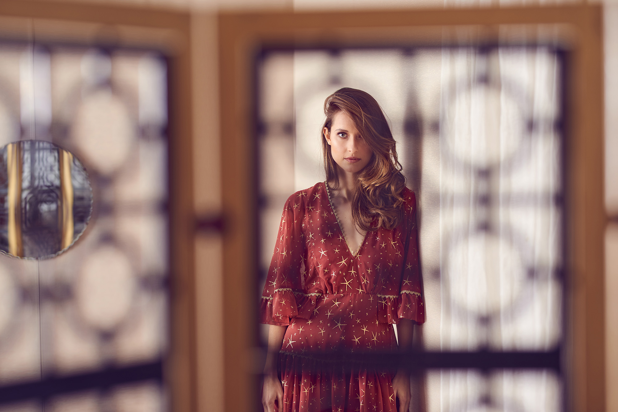Indoor shot of a fair-skinned adult woman in a star-patterned dress, posing in front of decorative glass panels with soft, diffused lighting.