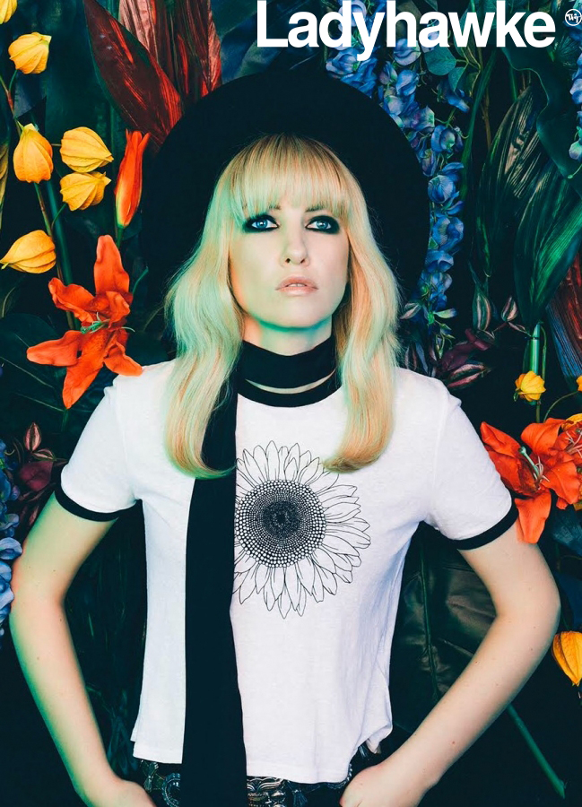 Blonde woman with bangs wearing a hat, scarf, and sunflower shirt stands against a vibrant floral backdrop in a portrait shot.
